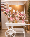 White wooden flower cart with canopy and large wheel displaying pink and white gift bouquets.