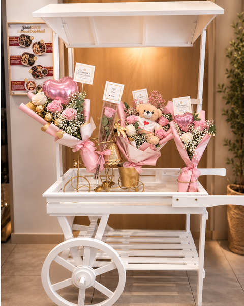 White wooden flower cart with canopy and large wheel displaying pink and white gift bouquets.