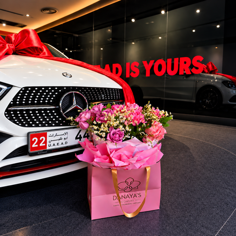 Pink rose bouquet in a Danaya’s Flowers gift bag beside a white Mercedes decorated with red ribbon.
