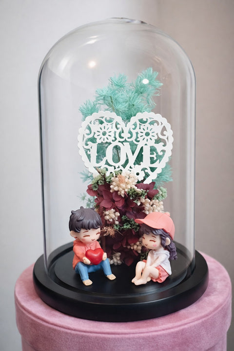 Decorative display with figurines under a glass dome featuring floral elements and a 'LOVE' heart.