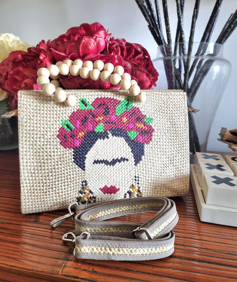 Woven handbag with Frida Kahlo design on a wooden surface with flowers and decorative items.