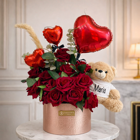 Bouquet of red roses with heart-shaped balloons and a teddy bear wearing a 'Maria' shirt on a marble surface.