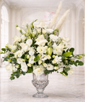 Luxury white floral arrangement in a crystal vase featuring white roses, lilies, lisianthus, eucalyptus, and pampas grass on a white marble background.