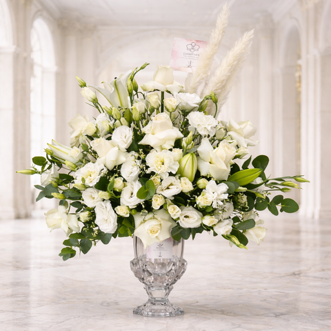 Luxury white floral arrangement in a crystal vase featuring white roses, lilies, lisianthus, eucalyptus, and pampas grass on a white marble background.
